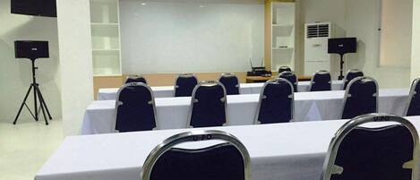 Une salle de conférence avec des rangées de tables et de chaises, un projecteur et un tableau blanc.