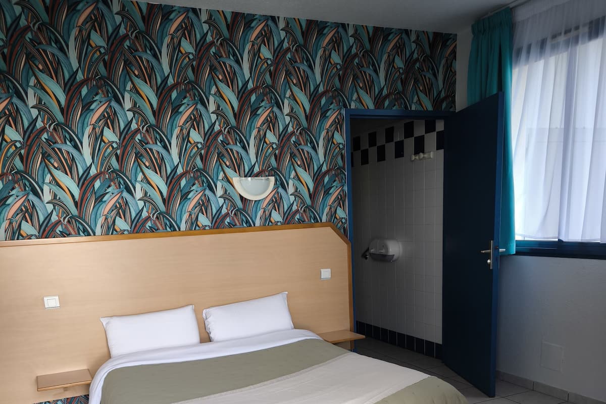 Une chambre d’hôtel comprenant un lit, une table de chevet, une salle de bain avec des toilettes et une fenêtre avec des rideaux.