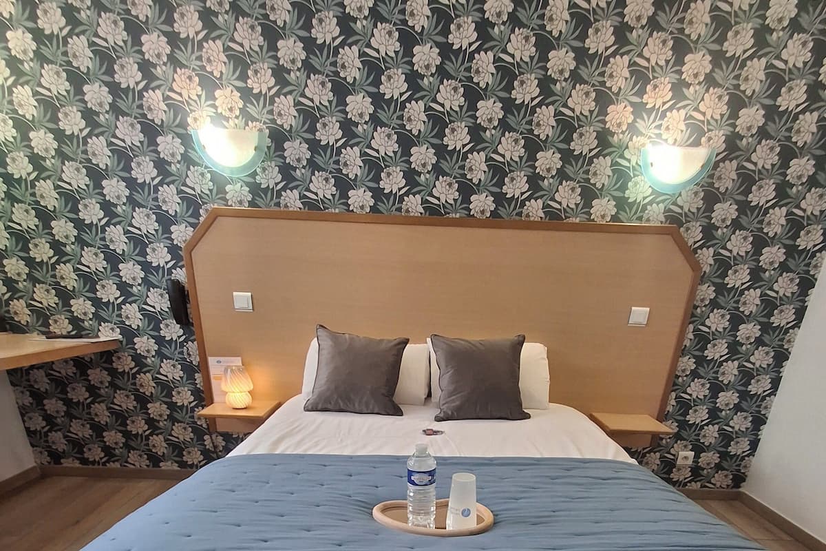 Une chambre d’hôtel avec un grand lit, deux oreillers, une table de chevet avec une lampe et un plateau avec une bouteille et une tasse.