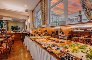 A buffet setup with various food items and a view of a sunset through large windows.