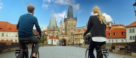 Two people riding bicycles on a cobblestone bridge with a historic building in the background.