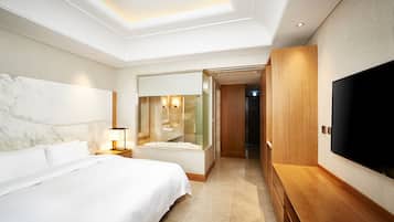 A modern hotel room with a large bed, a TV, a bathroom with a mirror, and a wooden bench.