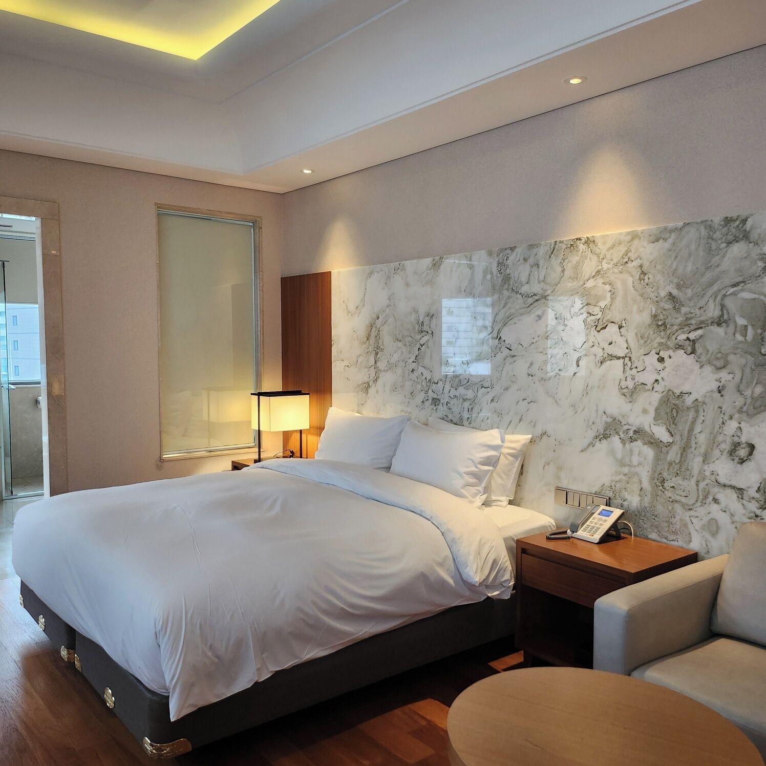 A modern hotel room with a large bed, a bedside table with a lamp, a marble wall, and a small round table.