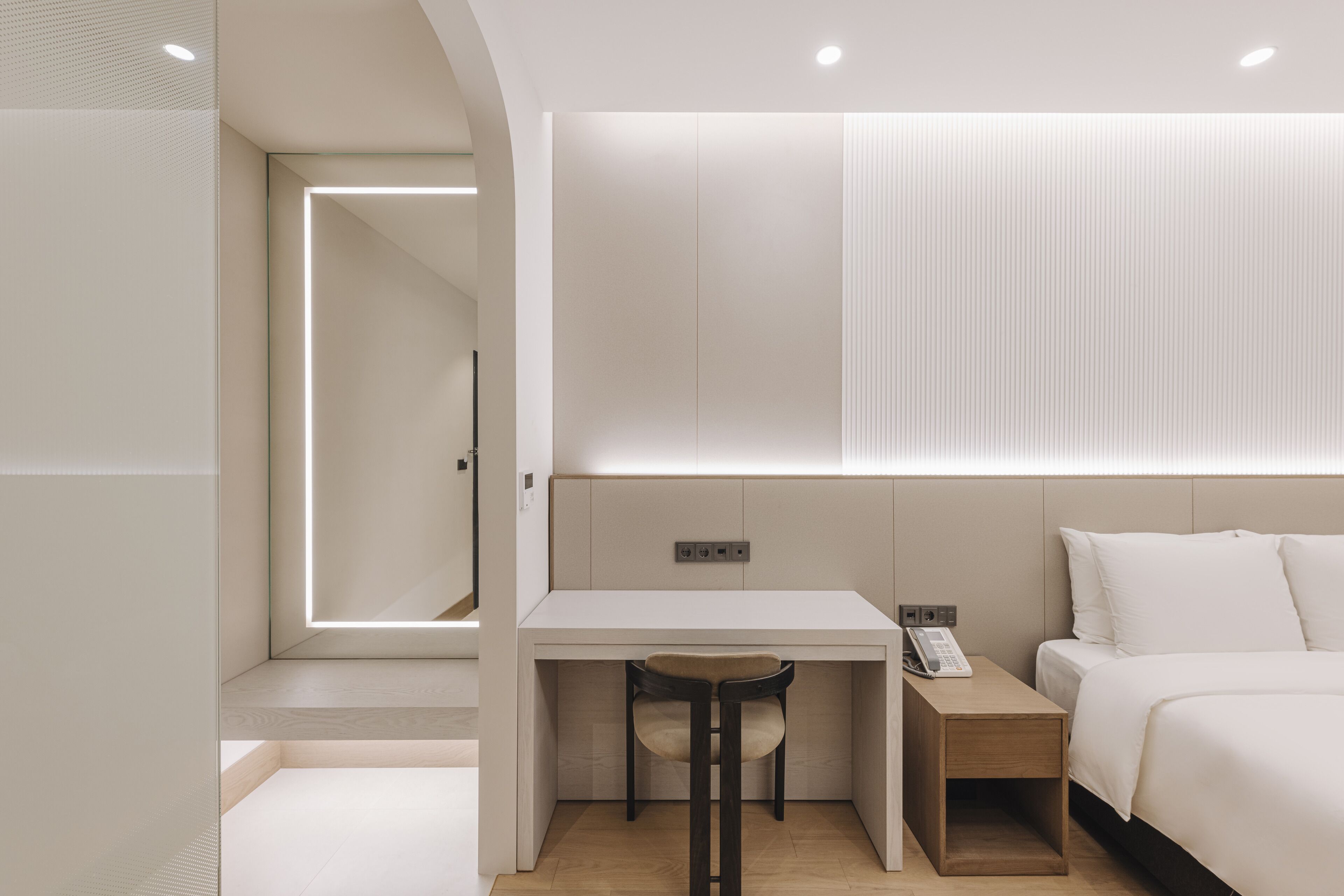 A modern hotel room with a minimalist design, featuring a bed, a desk with a chair, a bedside table, and a wall-mounted panel.