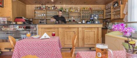 Un restaurante con interiores de madera, una zona de bar con estantes llenos de botellas y vasos, y una mesa con un jarro de cerveza y un florero.
