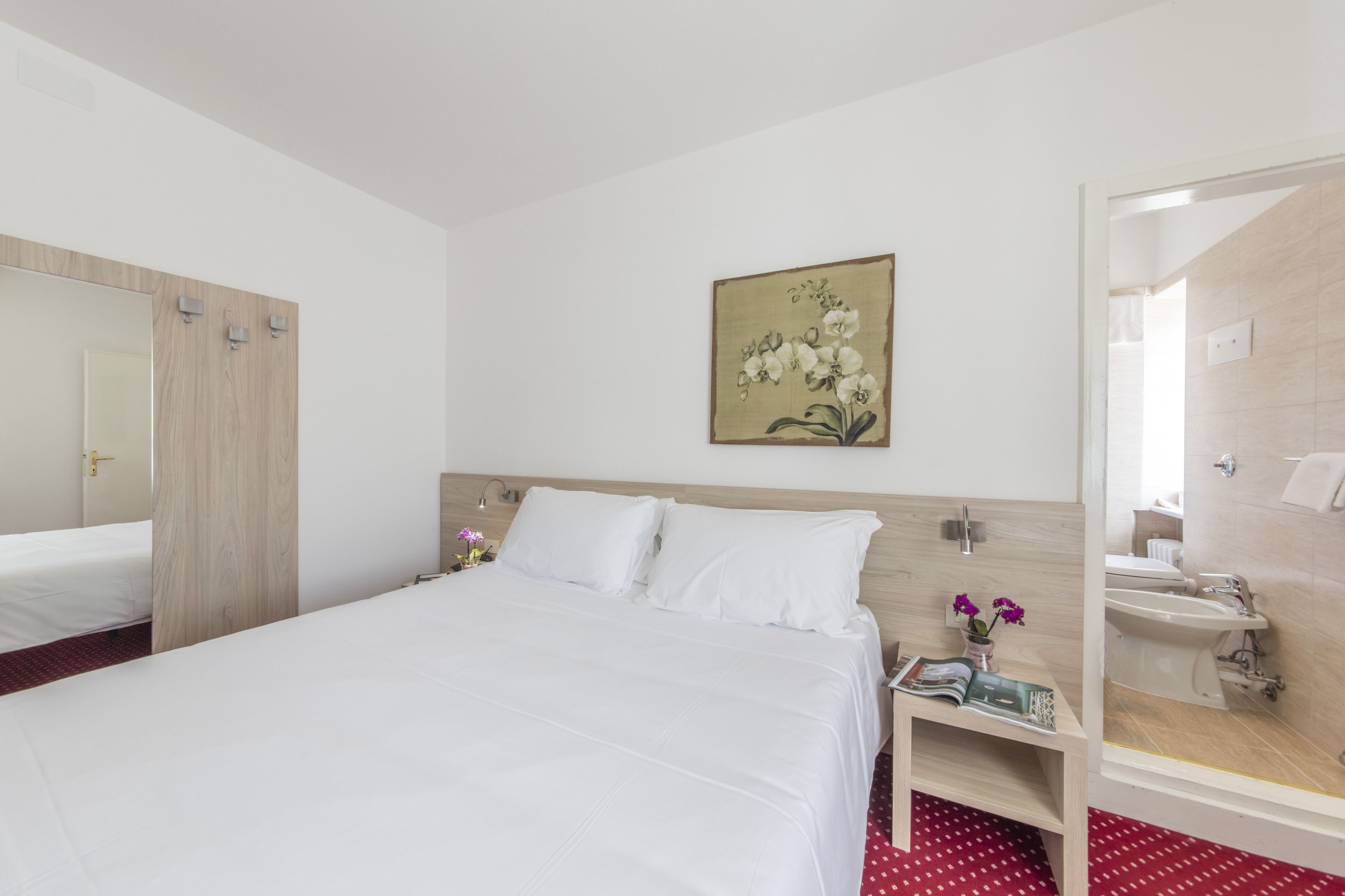 Una habitación de hotel con cama, una mesita de noche con una revista, un cuadro de flores en la pared y un baño con lavabo y bidé.