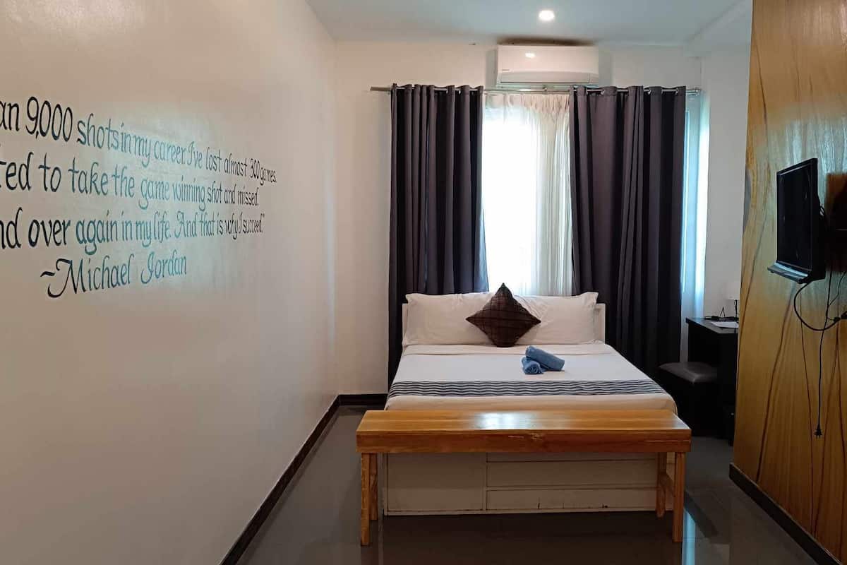 A hotel room with a bed, a wooden bench, a TV, and a wall with a motivational quote.