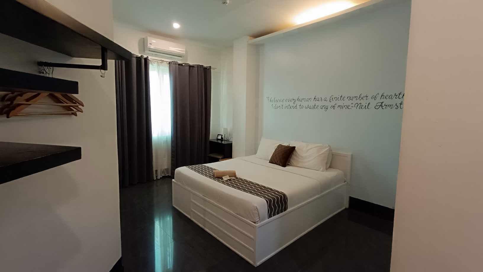 Una habitación de hotel moderna con una cama grande, una mesita de noche, aire acondicionado instalado en la pared y una cita en la pared.
