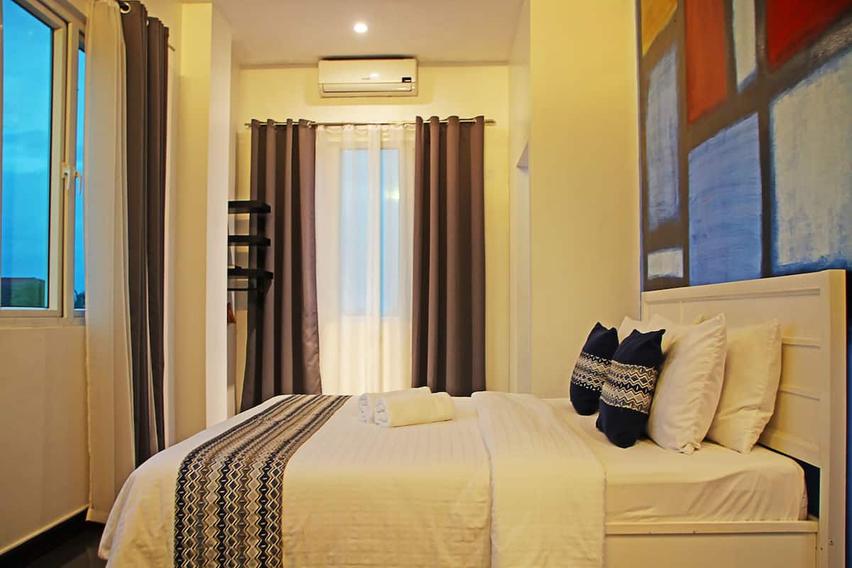 A hotel room with a bed, curtains, an air conditioning unit, and a painting on the wall.