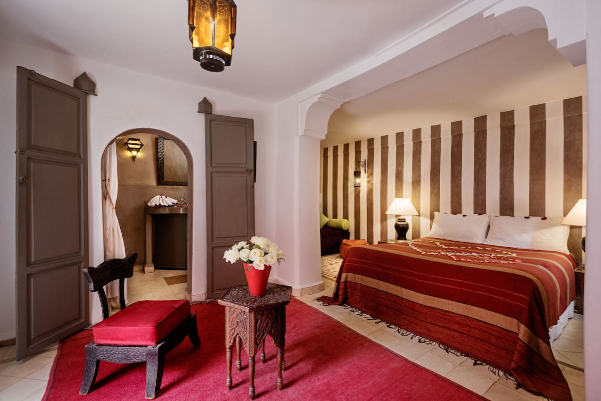 Une chambre d’hôtel avec un grand lit, un banc rouge, une petite table sur laquelle est posé un vase de fleurs, et une porte en arc donnant sur une autre pièce.