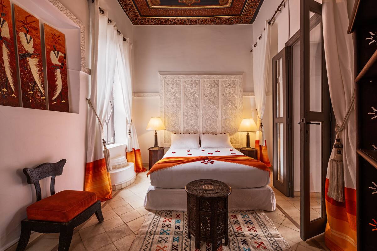 Une chambre d’hôtel avec un grand lit, deux tables de chevet avec des lampes, une petite table ronde, une chaise et un panneau décoratif mural.
