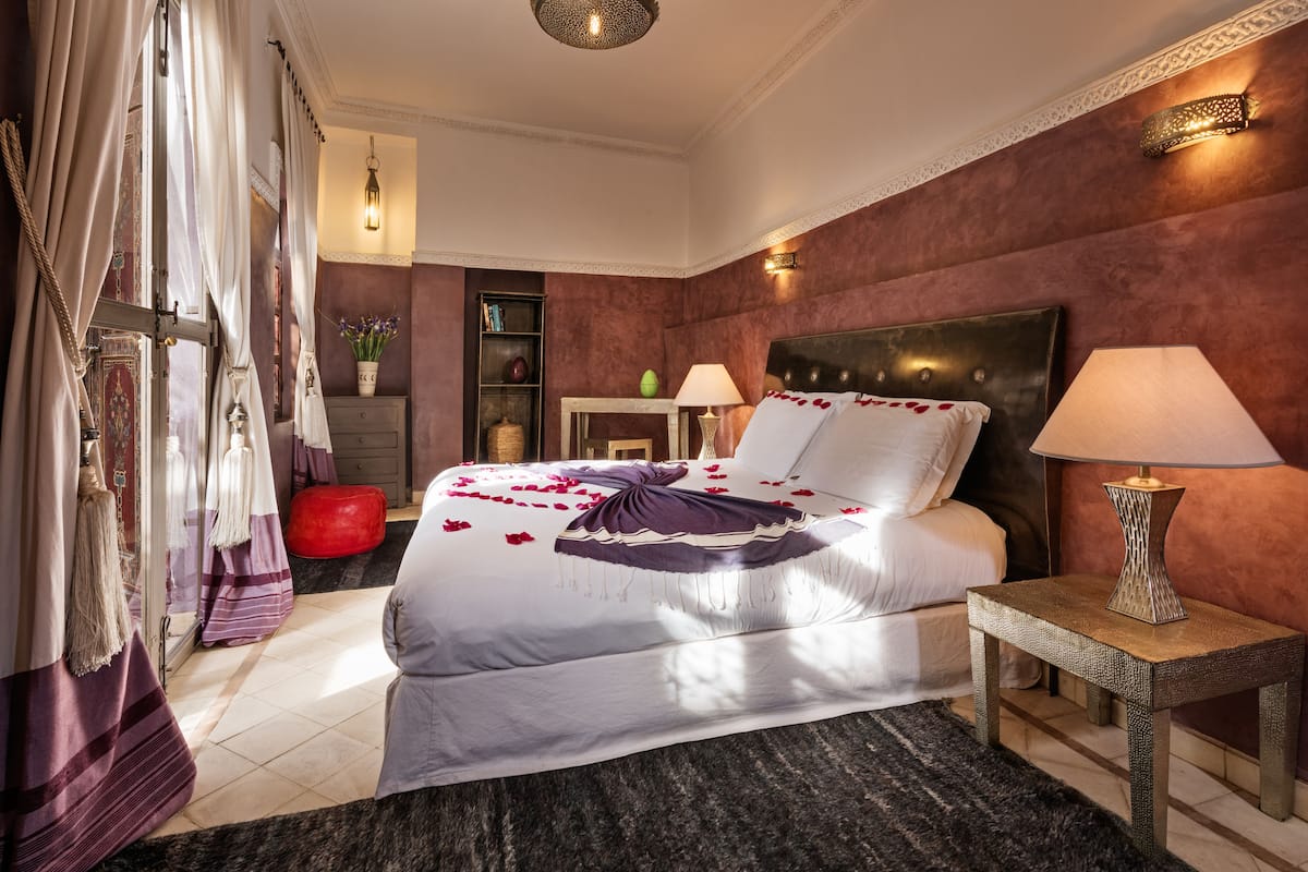 Une chambre d’hôtel élégamment aménagée, avec un grand lit, une table de chevet avec une lampe, une petite table d’appoint, une applique murale décorative et une étagère avec des livres et un vase.