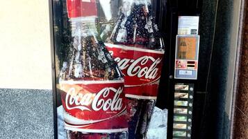A Coca-Cola vending machine with a large Coca-Cola bottle graphic on the front, a coin slot, and a display screen.