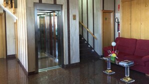A lobby with a glass elevator, a staircase, a red sofa, and a small table with magazines and flowers. - Hotel La Unión (Humanes de Madrid)