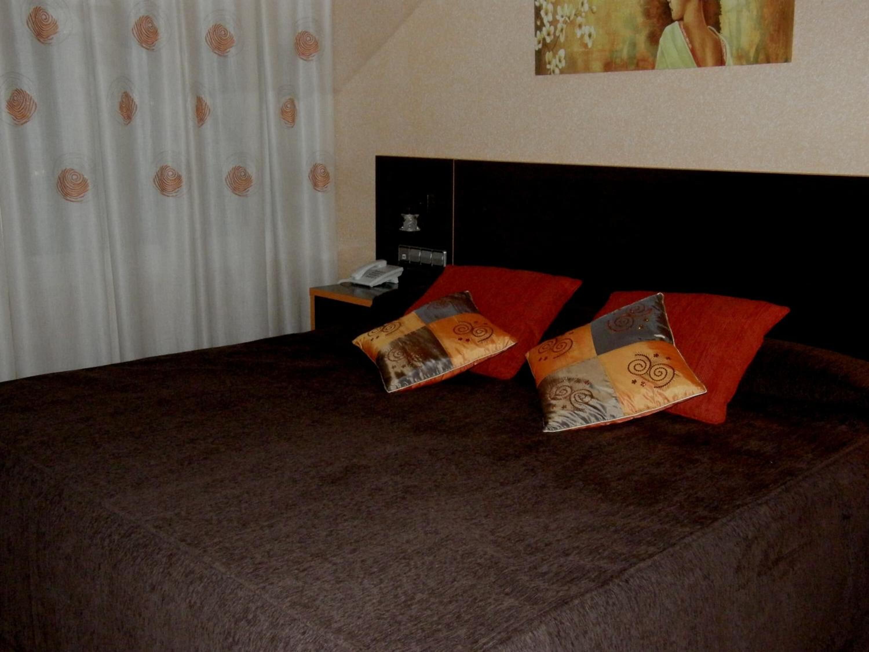 A hotel room with a bed, two red pillows, a nightstand with a phone, and a painting on the wall.