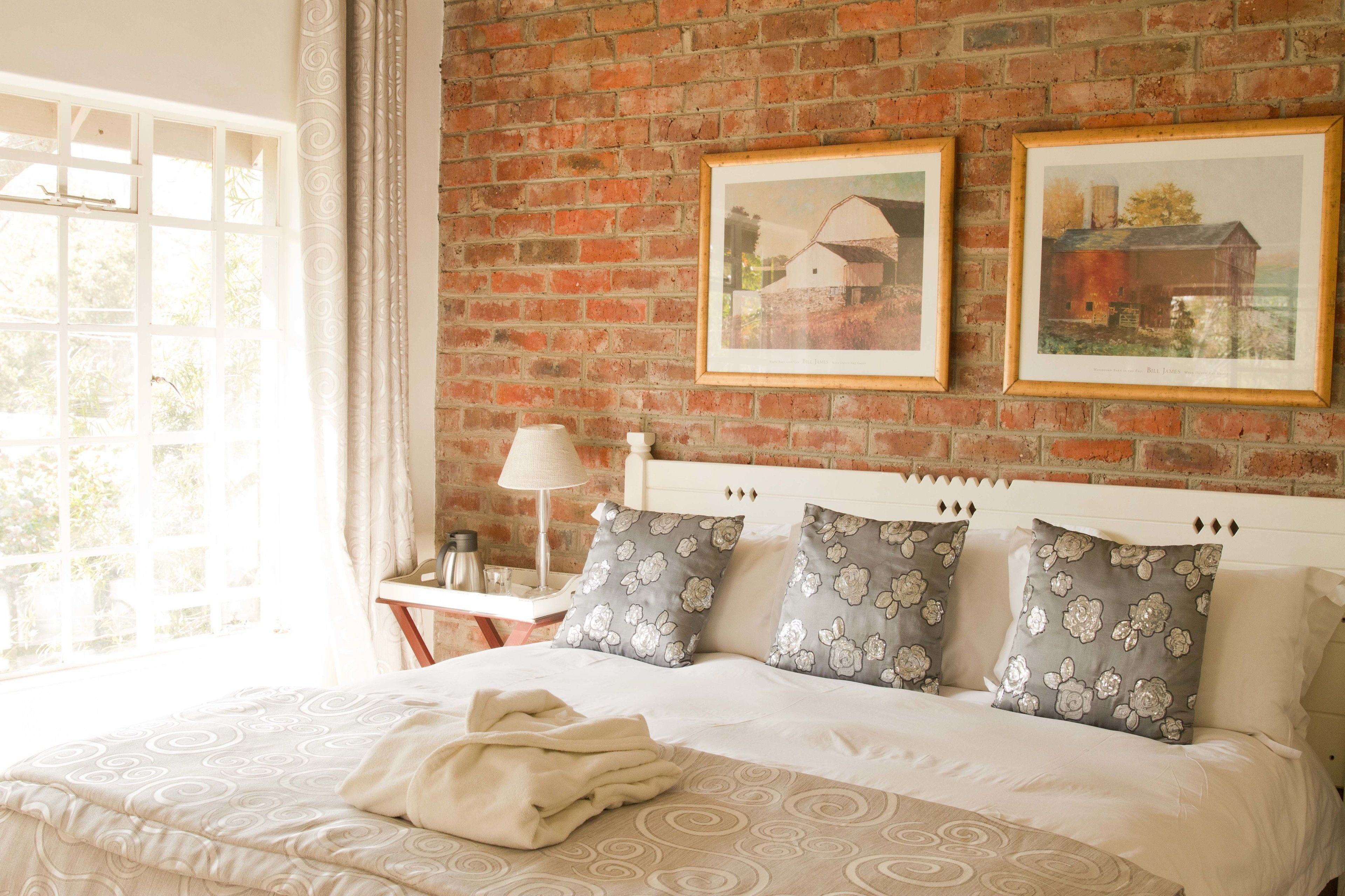 A bedroom with a brick wall, a bed with floral-patterned pillows, a bedside table with a lamp, and framed pictures on the wall.