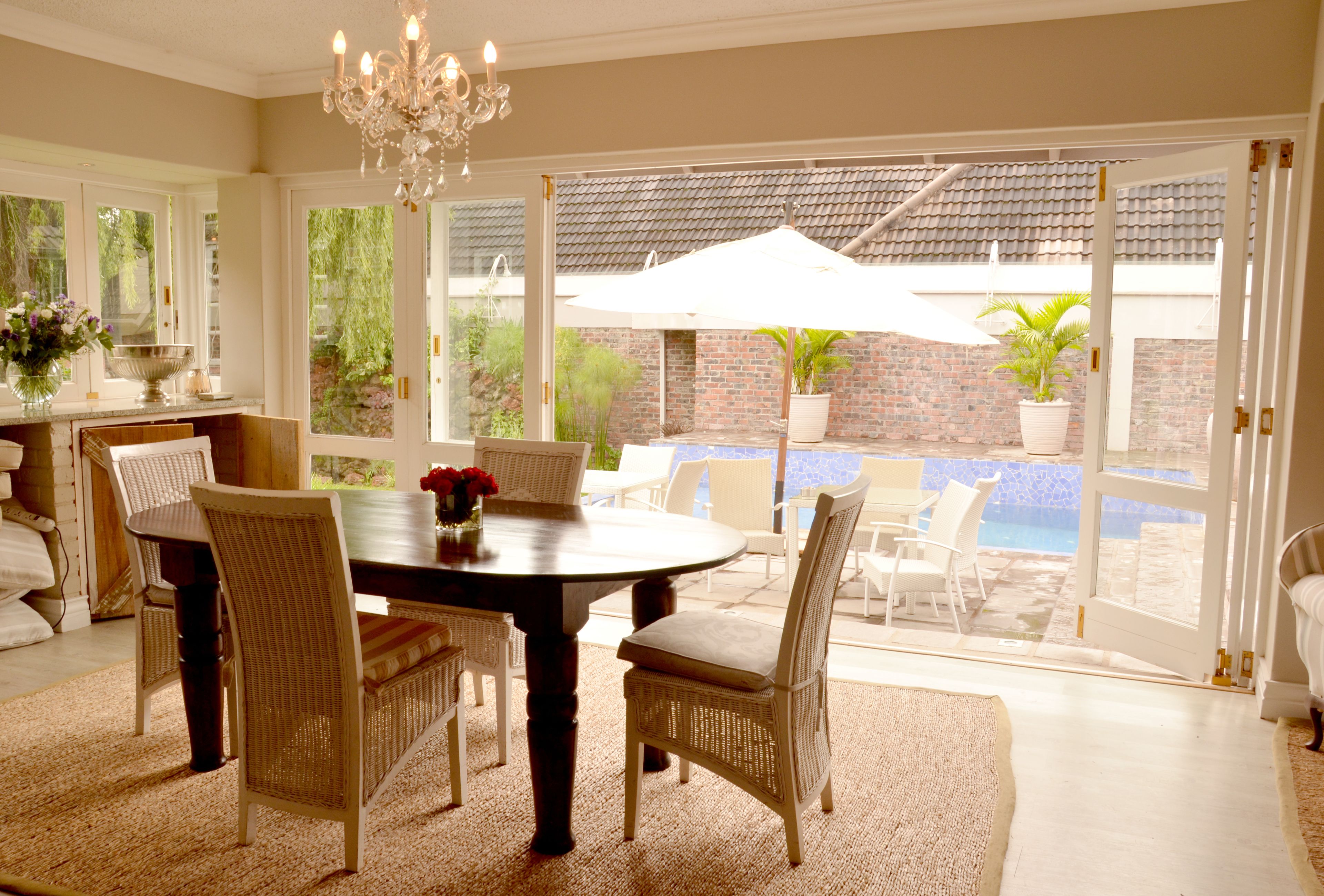 A dining area with a round table, wicker chairs, and a chandelier. There is a view of a swimming pool and a brick wall through large glass doors.