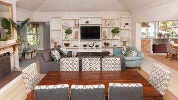 A spacious living room with a large wooden dining table, comfortable seating, a flat-screen TV, and a well-organized bookshelf.