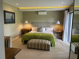A neatly arranged hotel room with a large bed, a green blanket, striped ottoman, bedside tables, and a painting on the wall.