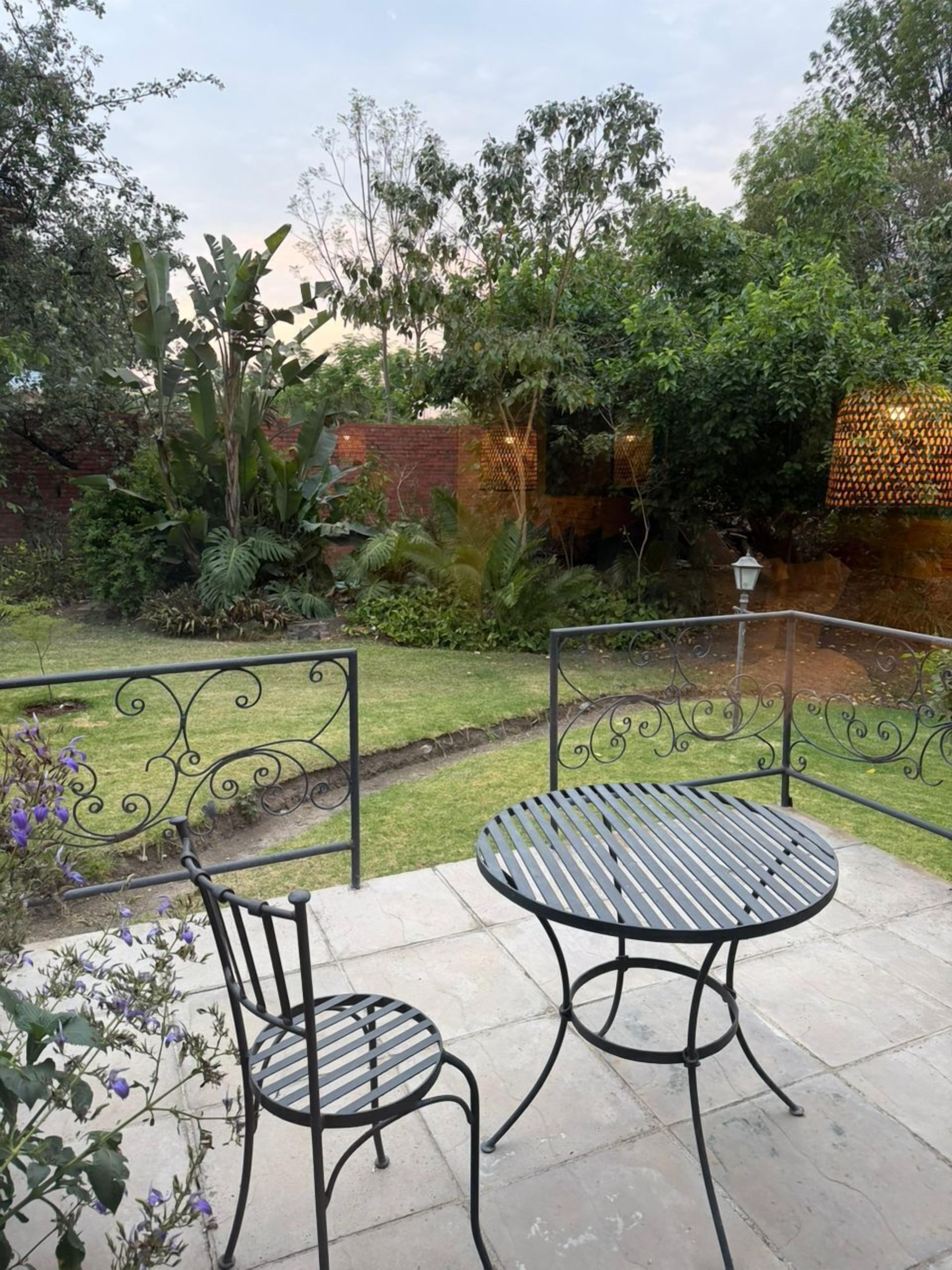 Een terras met een ronde tafel en twee stoelen, omringd door een metalen hek en weelderige beplanting.