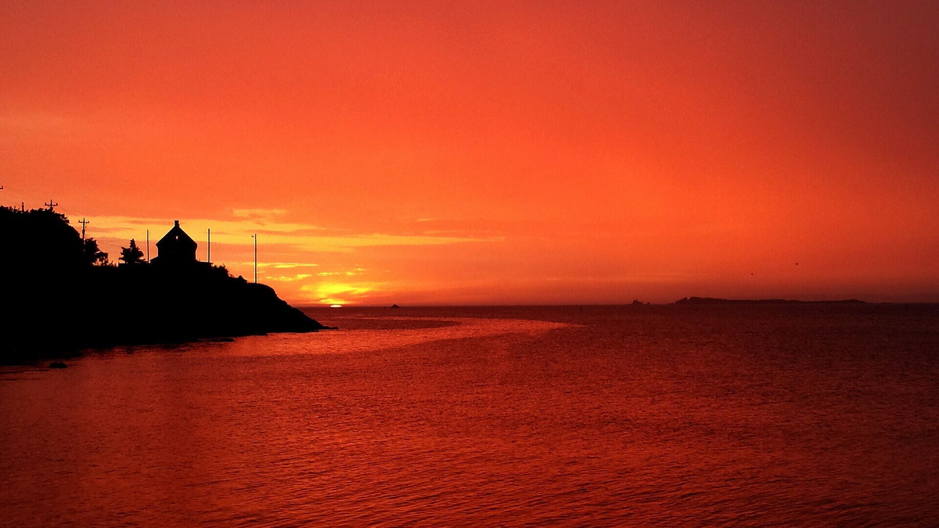 A coastal sunset with silhouettes of buildings and a flagpole on a cliff.