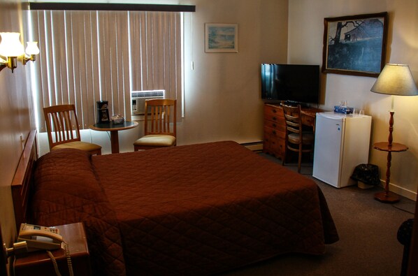 A hotel room with a large bed, a desk with a chair, a television, a refrigerator, and a lamp.