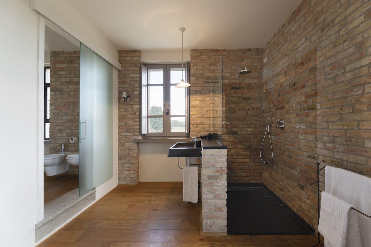 A modern bathroom with a brick wall, a wooden floor, a glass partition, a sink, a toilet, and a shower.