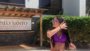 Een vrouw in een paars badpak en zwarte zwemshort die voor een bord staat met daarop de tekst Palo Santo Hotel Galapagos.