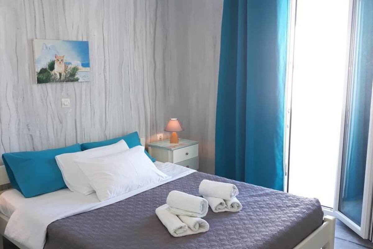 Una cama bien hecha con ropa de cama azul y blanca, un cuadro en la pared, una lámpara de noche y una ventana con cortinas azules.