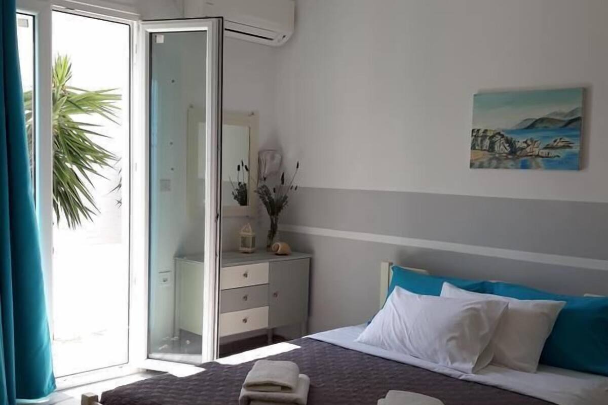 Un dormitorio con cama, mesita de noche, espejo, un cuadro y una ventana con cortinas.
