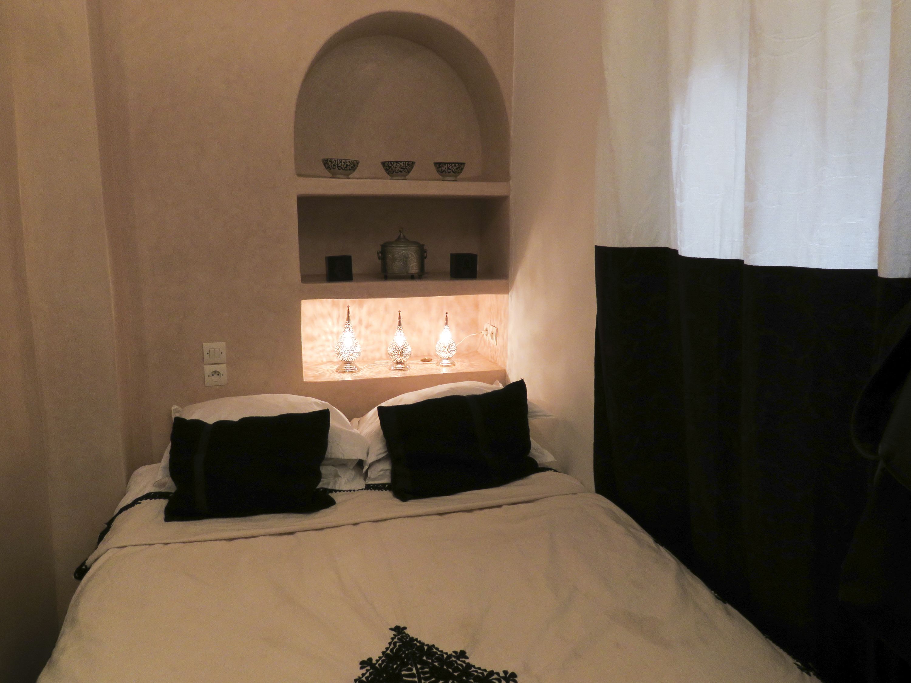 Ein Bett mit weißer Bettwäsche und schwarzen Kissen, ein Regal mit Dekoration und ein Fenster mit schwarzem Vorhang.