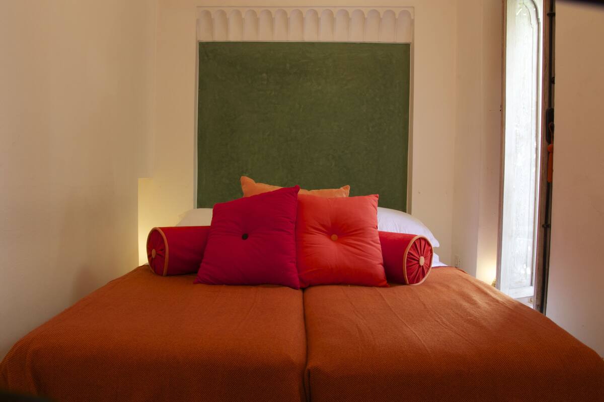 Een bed met twee oranje lakens en meerdere rode kussens.