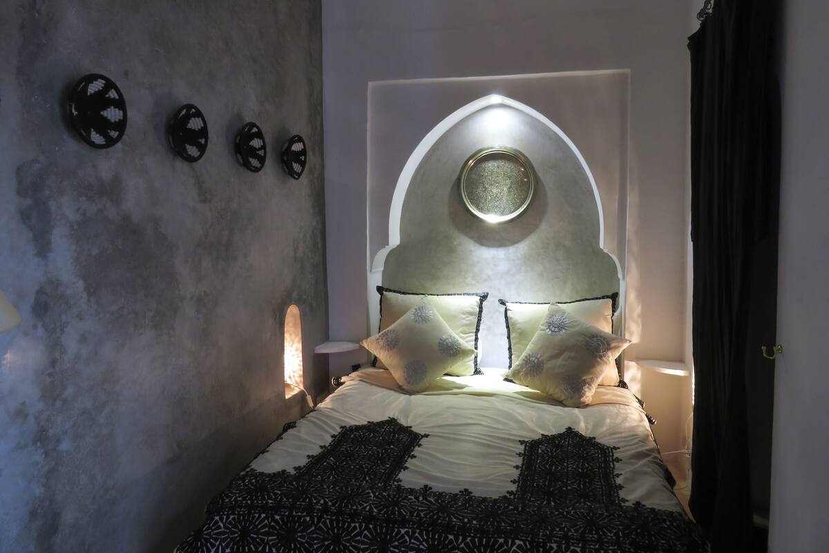 Een slaapkamer met een bed, een hangklok en een bijzonder, rondbogig raam.
