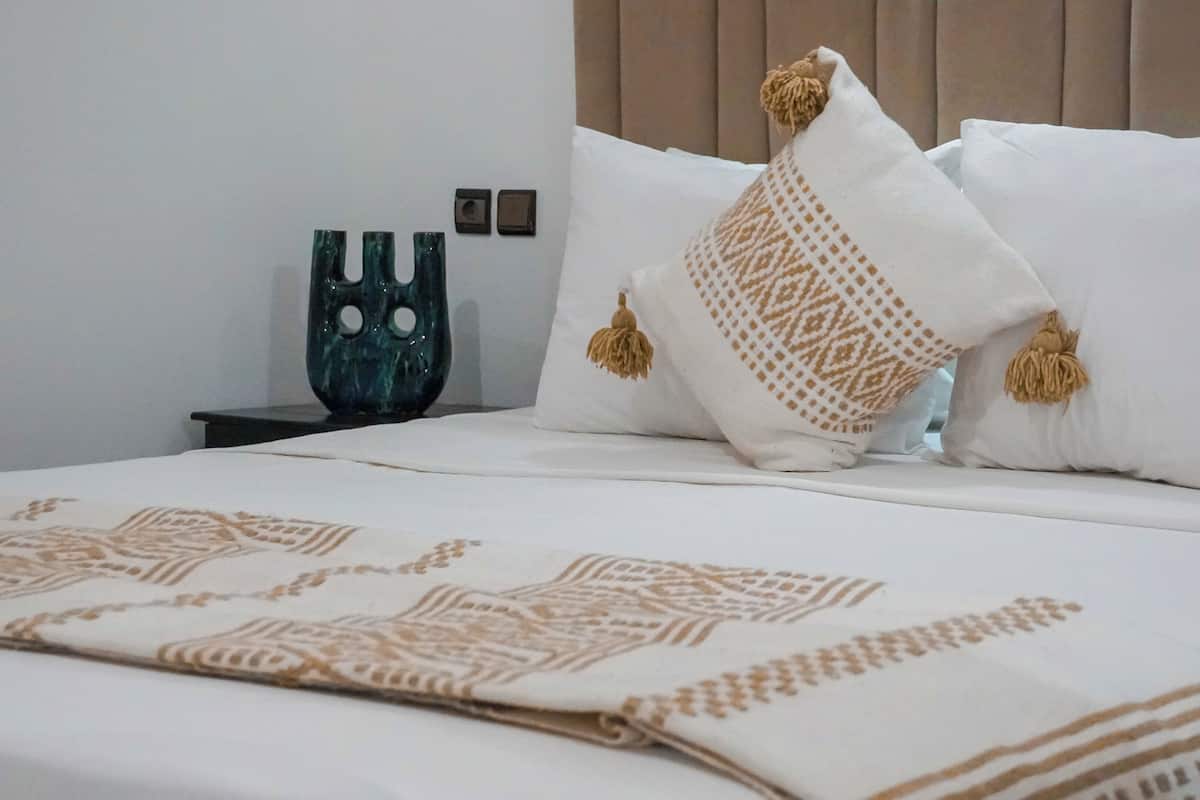Una cama bien hecha con ropa de cama estampada, un cojín decorativo y una mesita de noche con un florero.