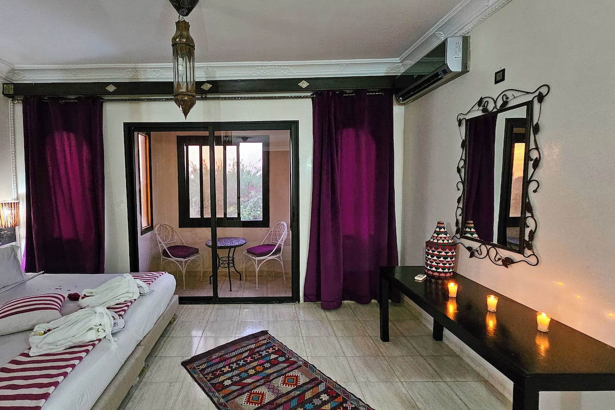 Habitación de hotel con cama, una mesita con velas, una silla y vistas a través de una ventana con cortinas.