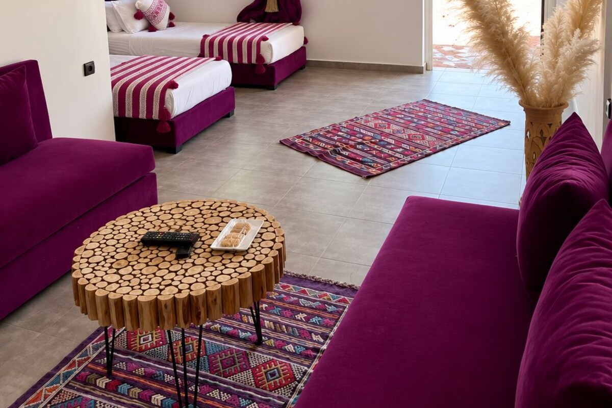 A room with two beds, a sofa, a round wooden table, and a view of greenery outside.