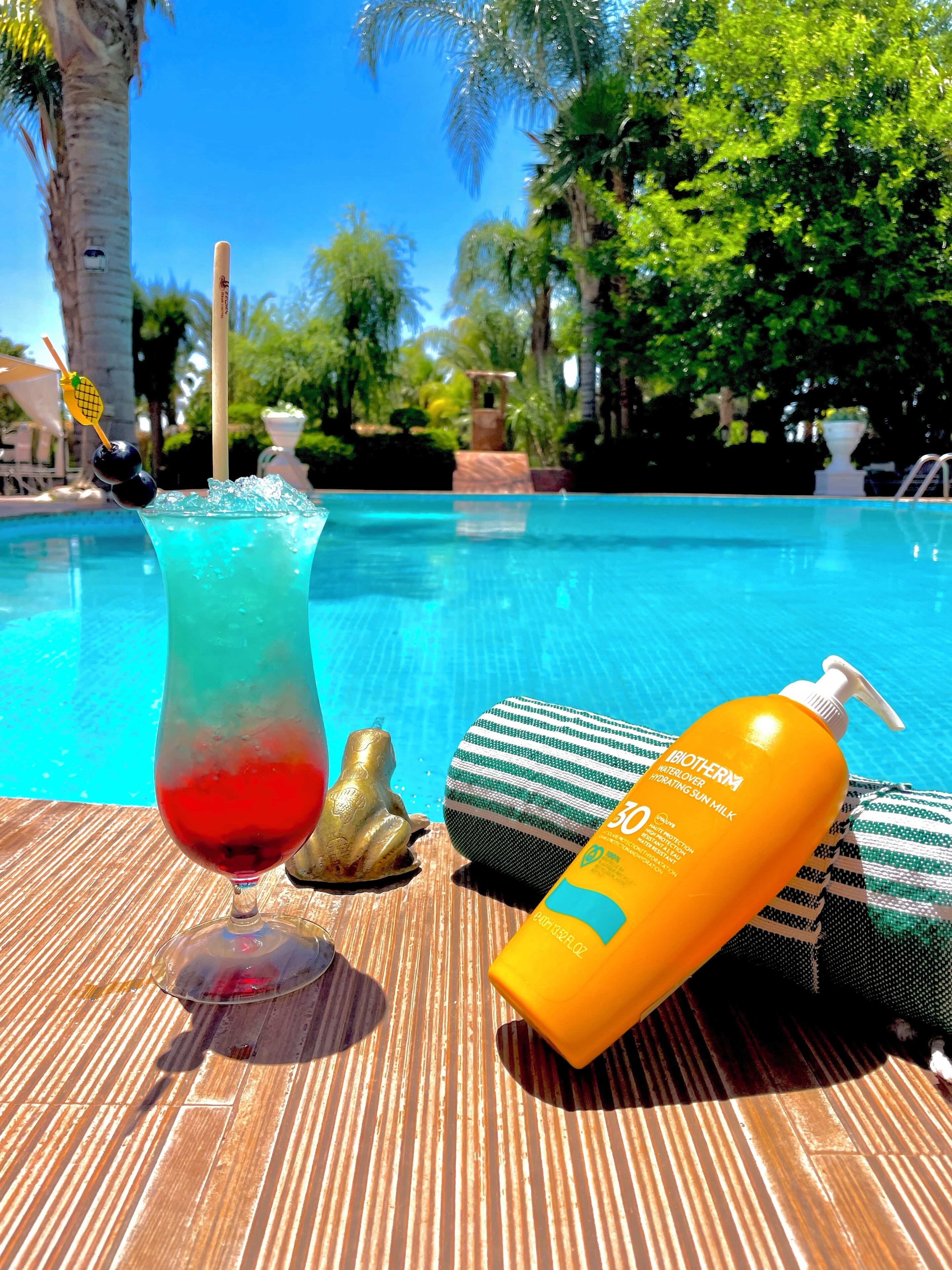 Ambiente à beira da piscina com uma bebida, um frasco de protetor solar e uma estátua de sapo decorativa.