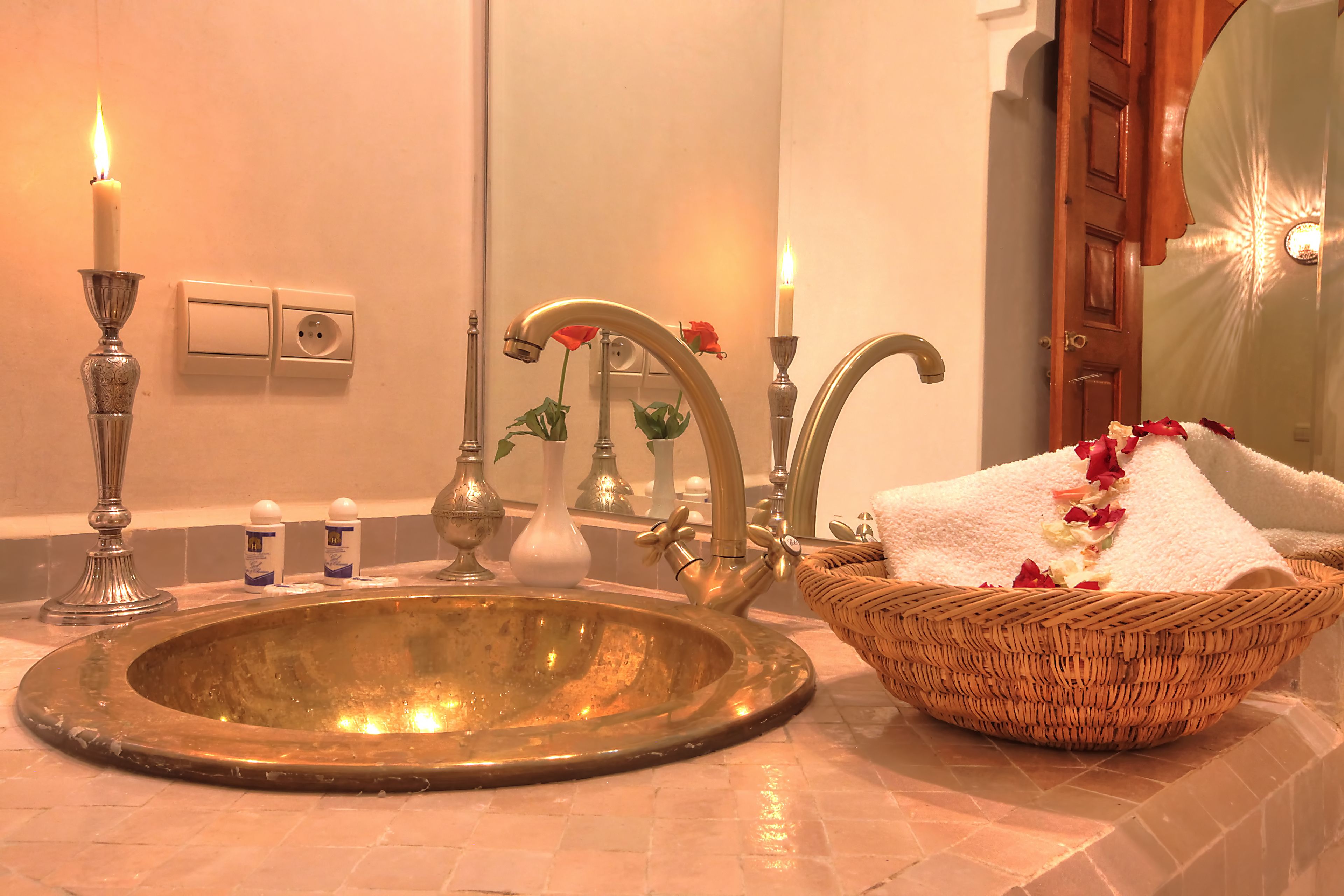 A bathroom with a gold sink, two lit candles, a mirror, a basket with a towel, and a vase with flowers.