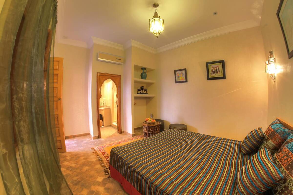 Un dormitorio con una cama grande, una puerta de madera, una mesita pequeña y una estantería con adornos.