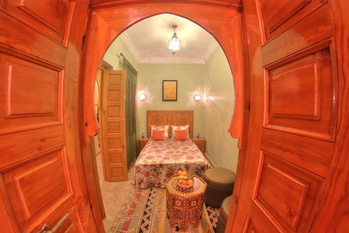 Una habitación con cama, mesita de noche, silla y lámpara de pared.