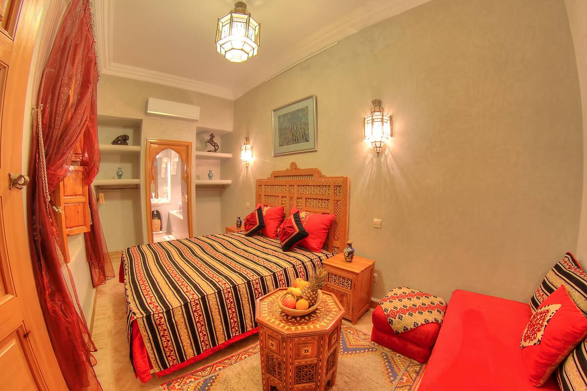 Un dormitorio con una cama grande, una mesita de noche de madera, una pequeña mesa con fruta y un sofá rojo.