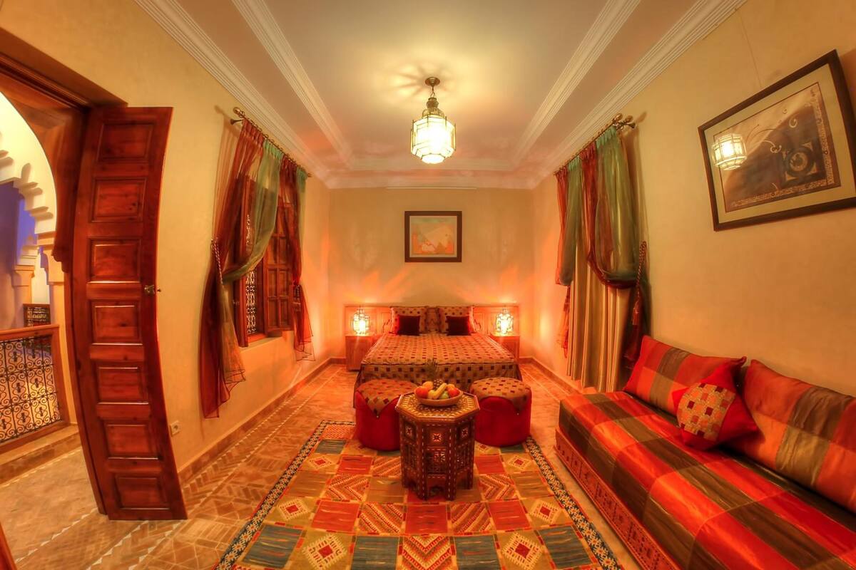 Una habitación tradicional con cama, sofá, una pequeña mesa con fruta, alfombras estampadas y cortinas.