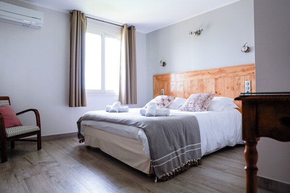 A bedroom with a wooden headboard, a bed with white bedding, a chair with a patterned cushion, and a window with curtains.