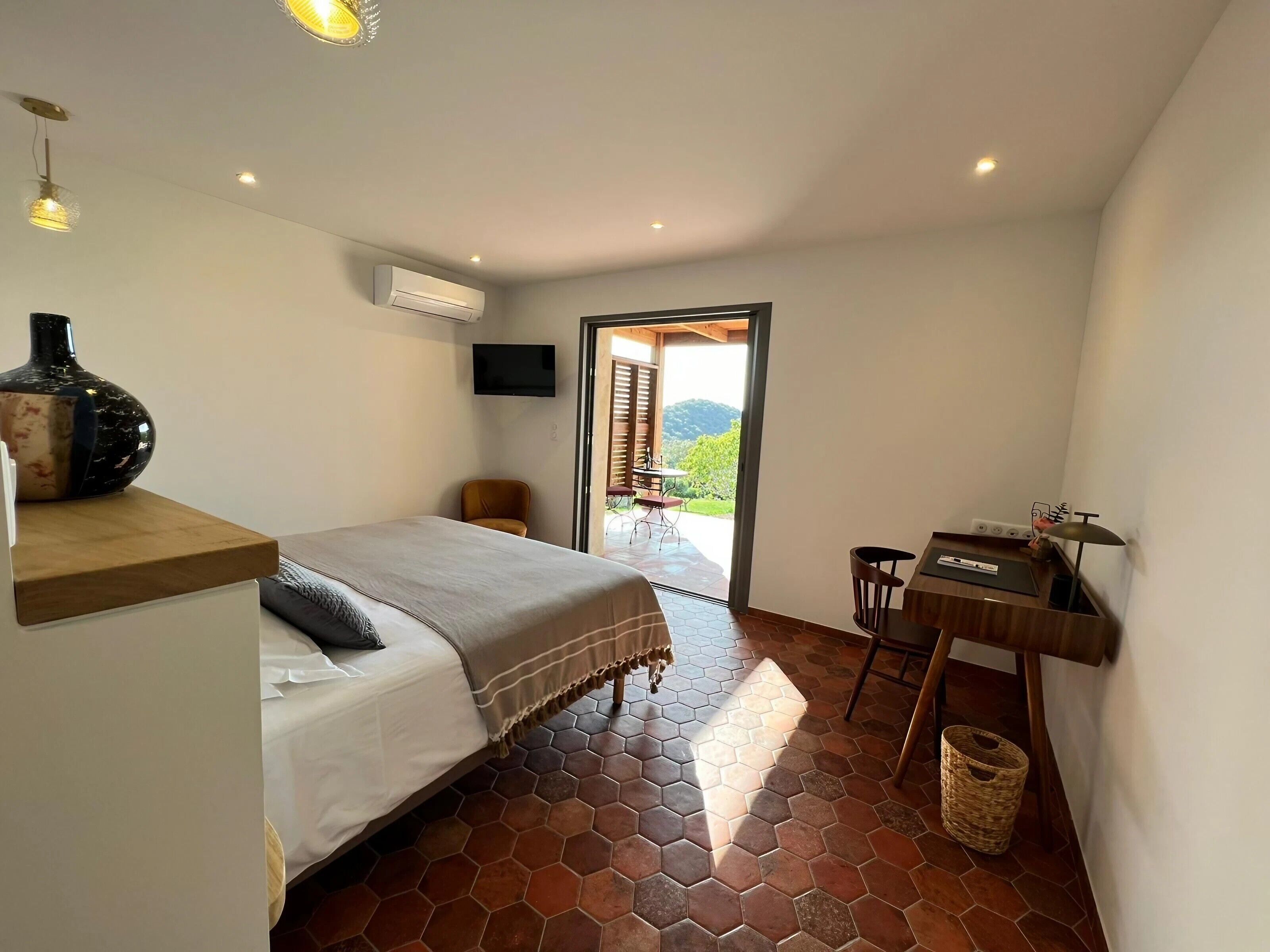 A bedroom with a bed, a desk, a chair, a television, and a view of the outdoors.