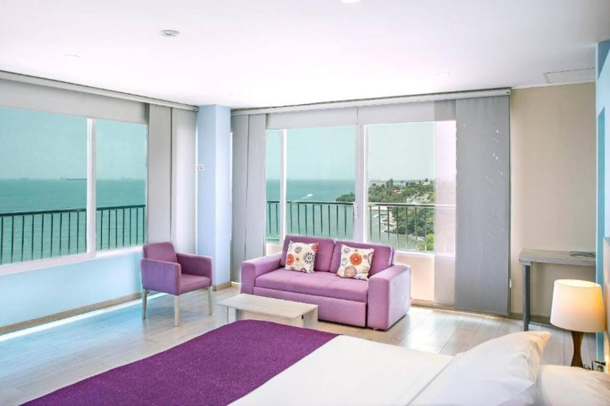 Une chambre d’hôtel moderne avec un grand lit, un canapé violet, un petit fauteuil et une vue sur la mer grâce à de grandes fenêtres.