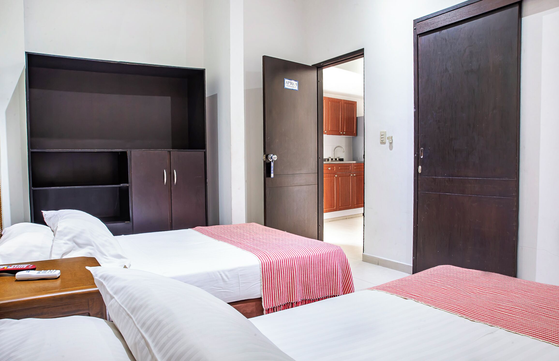 Habitación de hotel con dos camas, un armario, un escritorio con control remoto y una puerta que da a una zona de cocina.