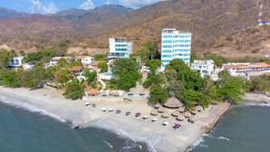 Vue aérienne d’une station balnéaire avec des villas en bord de mer, un bâtiment principal à plusieurs étages et une plage aménagée avec des parasols et des chaises longues.