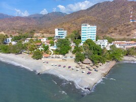 Vista aérea de un complejo turístico frente al mar, con villas en la playa, un edificio central de varios pisos y una zona de playa con sombrillas y sillones de descanso.