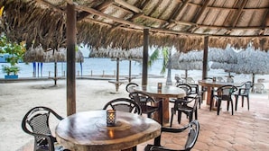 Un espace extérieur aménagé avec des parasols en chaume, des tables et des chaises en bois, offrant une vue sur une plage et la mer.