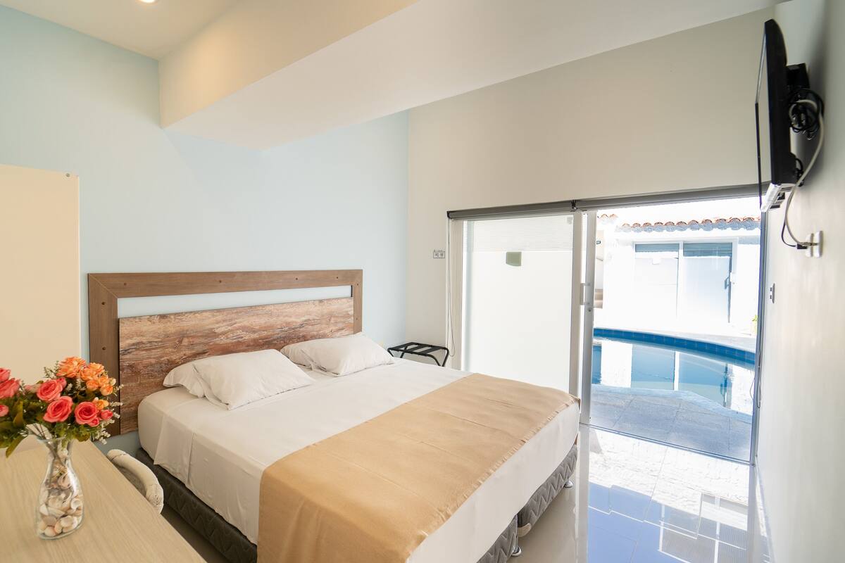 Une chambre à coucher avec un lit, une tête de lit en bois, une table de chevet ornée d’un vase de fleurs et une vue sur une piscine, visible à travers une porte coulissante.
